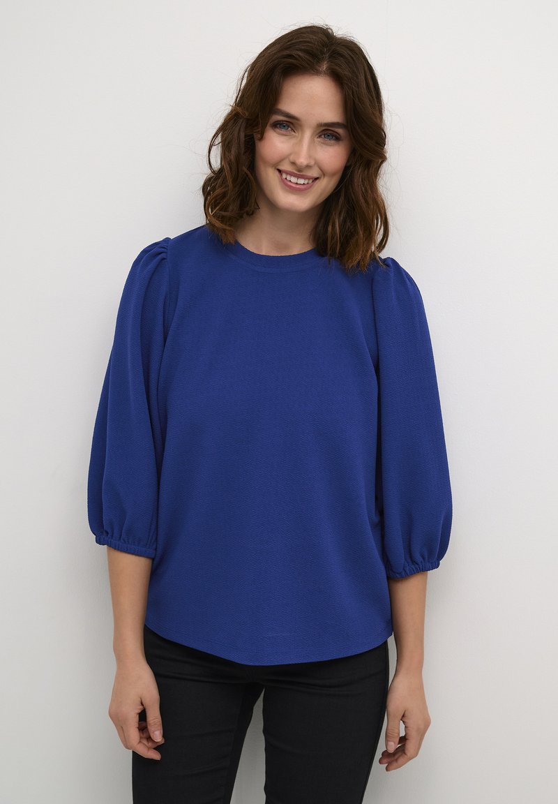 Kuningasinisest blouse'ist, millel on ümar kaelus, lühikesed puffvarrukad ja tekstureeritud kangas. Kaasa sobitatud tihedad mustad püksid.