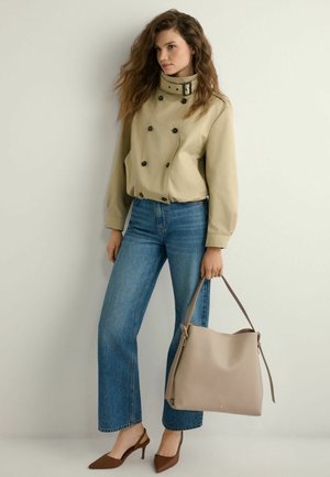 Kobieta w beżowej dwurzędowej kurtce, niebieskich dżinsach, brązowych szpiczastych szpilkach, trzymająca dużą beżową torebkę na tle jednolitej jasnej ściany.