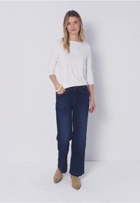 Žena stojící před bílým pozadím, oblečená v bílém topu s dlouhým rukávem, tmavých širokých džínách, béžových kotníkových botách a náramku na ruce.