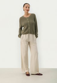 Olivgrüner gerippter Cardigan mit Knöpfen, kombiniert mit lockeren beigen Hosen mit Taschen und kastanienbraunen flachen Schuhen. Neutrale Hintergrund.