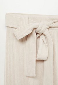 Pantalons en tissu côtelé beige avec une large taille et un détail de nœud devant. Texture douce avec des rayures verticales visibles.