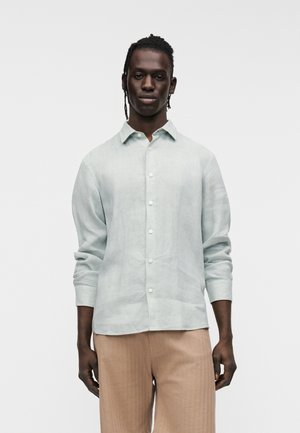 Camisa de lino azul claro con mangas largas, cierre de botones y cuello clásico, combinada con pantalones beige texturizados.