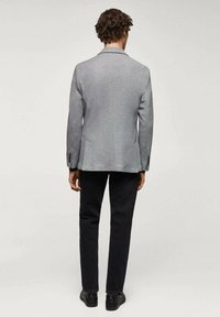 Blazer grigio con taglio classico, spacco singolo e due tasche anteriori, abbinato a pantaloni scuri e scarpe nere. Tessuto testurizzato con finitura liscia.