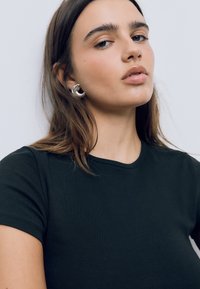 Černé tričko s krátkým rukávem a kulatým výstřihem. Model nosí stříbrné kruhové náušnice. Hladká textilie, vypasovaný střih, minimální detaily.