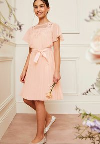 Abito plissettato rosa chiaro con un corpetto accentuato da pizzo e perline, maniche corte a volants e una cintura abbinata in raso. La indossatrice tiene un fiore.