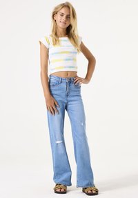 Crop top met meerkleurige horizontale strepen; high-waisted, lichtblauwe spijkerbroek met flared pijpen en beschadigde details; zwarte sandalen.