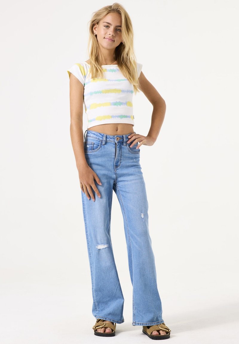 Crop top met meerkleurige horizontale strepen; high-waisted, lichtblauwe spijkerbroek met flared pijpen en beschadigde details; zwarte sandalen.