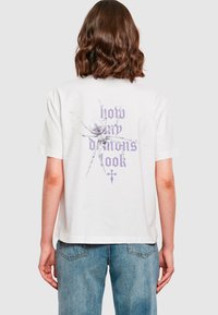 Balta medvilninė marškinėlių su grafika, vaizduojančia vorą, ir violetiniu tekstu "laikyk mano demonus" ant nugaros. Trumpomis rankovėmis, laisvas kirpimas.