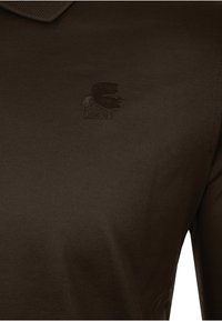 Nahaufnahme eines dunkelbraunen Poloshirts mit einem kleinen, ton-in-ton gestickten Logo eines Männerprofils, das Sonnenbrillen trägt und einen Pferdeschwanz hat.