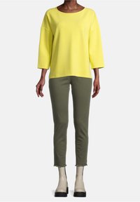 Geel, langarm shirt met een wijde halslijn; gecombineerd met slim-fit, olijfgroene broek en beige enkellaarsjes met zwarte accenten.