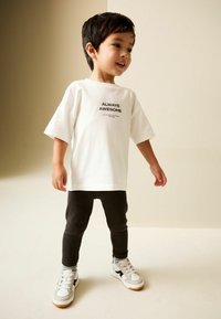 Tricou alb cu mâneci scurte, cu textul "ÎNTOTDEAUNA FENOMENAL" imprimat în negru, asortat cu colanți negri și adidași albi cu accente negre.