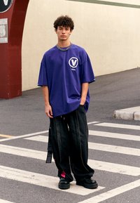 Camiseta de algodón púrpura de gran tamaño con un logo circular, combinada con pantalones de mezclilla negros holgados con un acento rosa, llevados con botas negras gruesas.