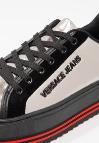 Baskets plateformes noires et argent métallisé avec lacets noirs, ornées du logo "VERSACE JEANS" et d'une semelle rayée rouge. Matériaux en velours doux et brillants.