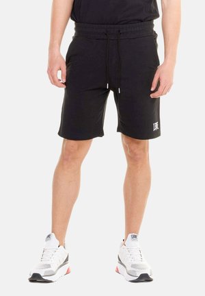 Man draagt zwarte korte broek met trekkoorden en witte sportschoenen, staand tegen een effen achtergrond.