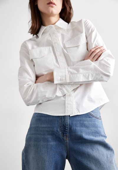 Cămașă albă cu nasturi, prevăzută cu două buzunare pe piept, asociată cu blugi denim de culoare albastră deschisă. Cămașa are un guler structurat și mâneci lungi.