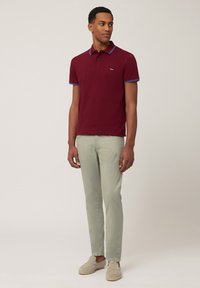 Burgundska polo majica s plavim detaljima, uskog kroja. Uparen s svijetlozelenim hlačama i bež kliznim cipelama, model stoji protiv neutralne pozadine.