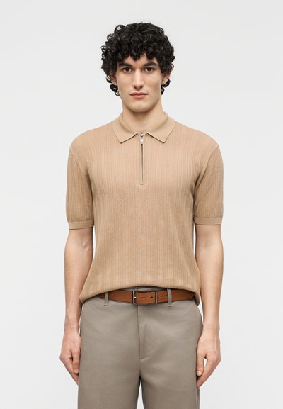 Homme aux cheveux bouclés portant un polo en maille beige à manches courtes, une ceinture marron et un pantalon gris, debout devant un fond uni.