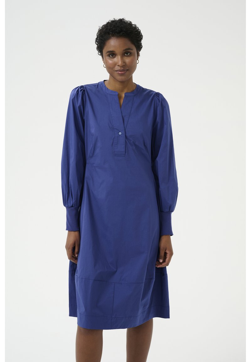 Izpūsts garais piedurkņu kleita karali zilo kokvilnā, ar V-veida kaklu un pogu detaļu pie apkakles, ar brīvu piegriezumu un apakšējo hem līniju zem ceļa.