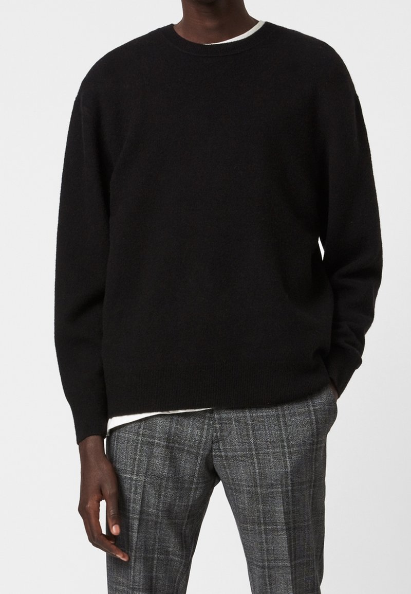 Schwarzer Strickpullover mit Rundhalsausschnitt, gerippten Bündchen und Saum, getragen über einem weißen Hemd, kombiniert mit grauen karierten Hosen.