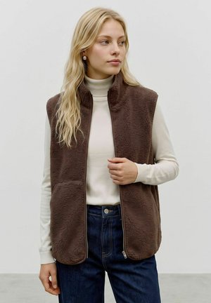 Femme blonde portant des boucles d'oreilles en perles, un col roulé blanc, un jean bleu foncé et un gilet polaire marron zippé, regardant sur le côté.