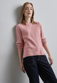 Hellrosa gestrickter Pullover mit gerippter Struktur, der ein diagonales Muster aufweist und einen Rundhalsausschnitt hat. Kombiniert mit dunkelblauen Jeans.