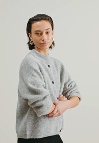 Harmaa neulecardigan, jossa on korkea kaulus, pitkät hihat ja mustat napit. Pehmeä rakenne ja rento malli, yhdistetty mustaan asusteeseen.