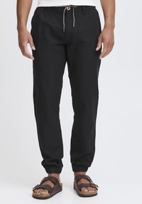 Zwarte drawstring broek van lichtgewicht stof met elastische boorden, voorzien van een houten knoop en contrasterende stiksels. Draag ze met bruine sandalen.