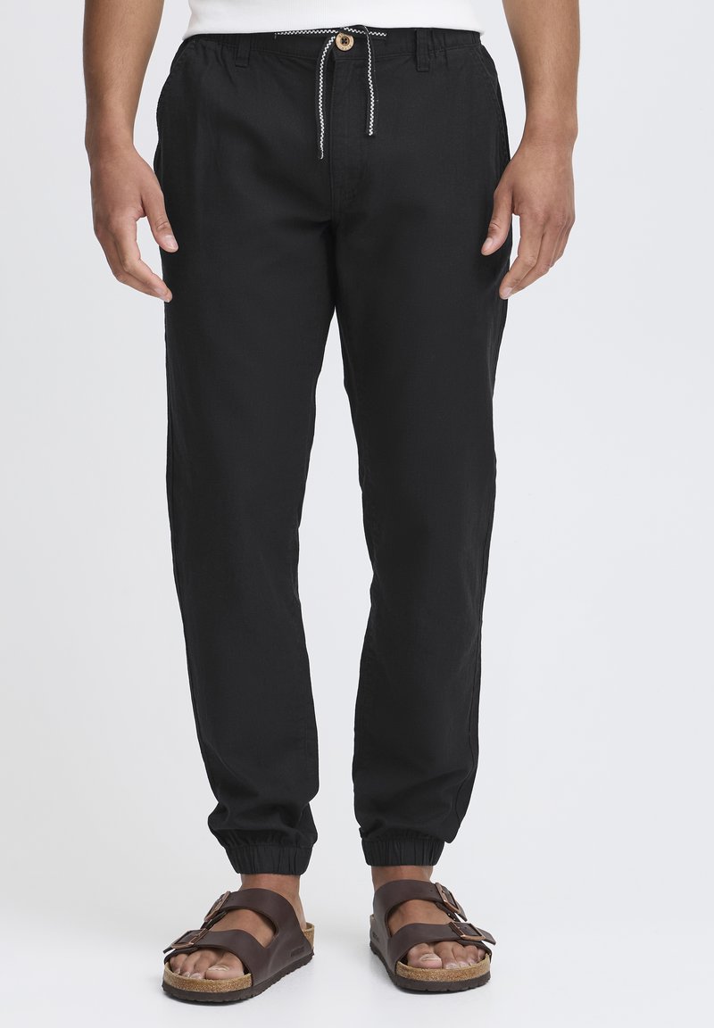 Zwarte drawstring broek van lichtgewicht stof met elastische boorden, voorzien van een houten knoop en contrasterende stiksels. Draag ze met bruine sandalen.