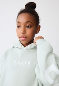 Mündi rohelise tooniga kapuutsiga dressipluus esiküljel logoga "BAKER BY TED BAKER." Omadused hõlmavad paela kapuutsi ja ülemõõdulisi varrukaid valge aktsendiga.
