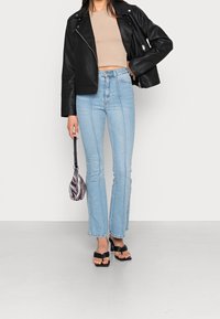 Svart lädercropped jacka, beige åtsittande topp, ljusblå utsvängda jeans med sömdekor, svarta plattformssandaler och en mönstrad handväska.