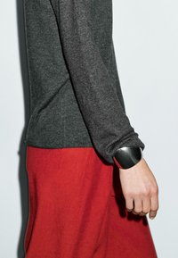 Un gros plan du profil latéral d'une personne portant une chemise à manches longues grise et un pantalon rouge, avec un bracelet en argent bien visible à son poignet.