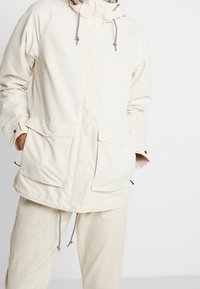 Osoba v krémově bílé mikině s kapucí, předními kapsami a stahovacími šňůrkami, v kombinaci s odpovídajícími krémově bílými kalhotami, proti jednobarevnému pozadí.