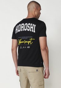 Camiseta negra de manga corta que presenta "KOROSHI" en grandes letras blancas en la parte posterior y acentos amarillos, con una textura de algodón suave.