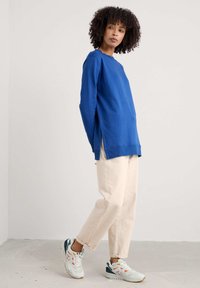 Kék, hosszúujjú pulóver, laza szabással, strukturált anyagból, oldalsó hasítékokkal, bézs, rövidnadrággal és színes sportcipőkkel párosítva.