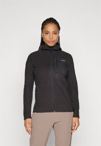 Woman wearing a black zip-up textured fleece jacket with a hood and chest pocket, paired with light brown pants, standing against a plain background.