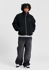 Musta fleece-tyyppinen vetoketjullinen takki, jossa on korkea kaulus, sivutaskut ja teksturoitu kangas. Yhdistetty löysien mustien housujen kanssa ja kontrastiväristen tennareiden kanssa.