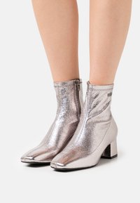 Botines metálicos plateados hasta el tobillo con puntera afilada y tacón cuadrado. Cuentan con una cremallera lateral y una textura lisa y brillante.