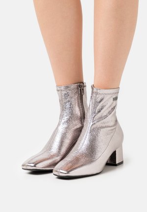Botines metálicos plateados hasta el tobillo con puntera afilada y tacón cuadrado. Cuentan con una cremallera lateral y una textura lisa y brillante.