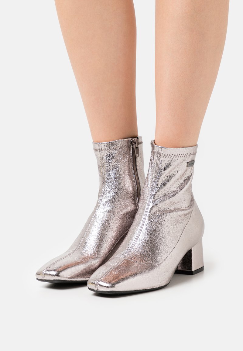 Botines metálicos plateados hasta el tobillo con puntera afilada y tacón cuadrado. Cuentan con una cremallera lateral y una textura lisa y brillante.