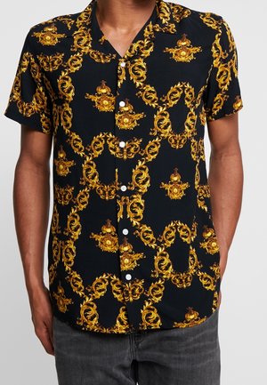 Camisa negra de manga corta abotonada con un elaborado patrón barroco en amarillo y dorado, usada por una persona con vaqueros oscuros.