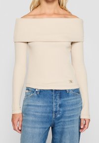 Frau in einem beigen, schulterfreien Langarmtop, kombiniert mit blauen, hoch taillierten Jeans, die Hände entspannt an den Seiten.