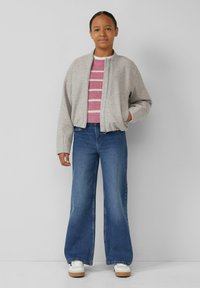 Giubbotto bomber grigio chiaro con zipper frontale, maglione a righe rosa, jeans a gamba larga blu e sneaker bianche con suole color beige.