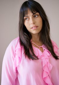 Femme aux longs cheveux noirs portant une blouse rose à volants, regardant légèrement sur le côté devant un fond neutre.