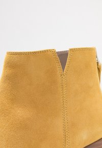 Botín amarillo de ante hasta el tobillo con un diseño de caña partida, textura suave y costuras visibles a lo largo de los bordes. Punta cerrada y minimalismo en los herrajes.