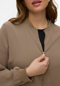 Veste bomber marron avec col et poignets côtelés, dotée d'une fermeture éclair dorée. Tissu doux, manches légèrement bouffantes et coupe ajustée.