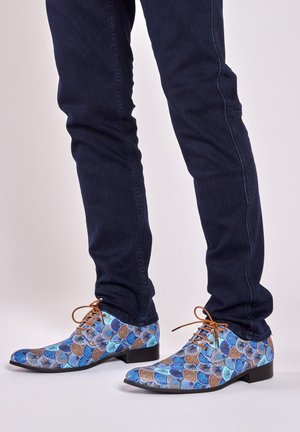 Blauwe en bruine gestreepte dresschoenen met een spitse neus en platte hak; voorzien van textielmateriaal en bruine veters. Draagt bij donkere jeans.