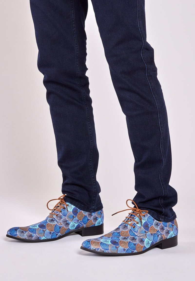 Blue and brown patterned dress shoes with a pointed toe and flat heel; features textured fabric and brown laces. Worn with dark jeans.