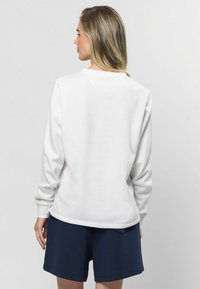 Biela mikina s dlhými rukávmi, okrúhlym výstrihom a rebrovanými manžetami; skombinovaná s námorníckymi šortkami. Hladká textúra, neformálny dizajn, minimálne detaily.