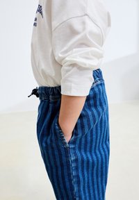 Pantaloni din denim cu dungi albastre, cu talie elastică, având buzunare laterale și șnur. Asortați cu o cămașă albă cu mâneci lungi.
