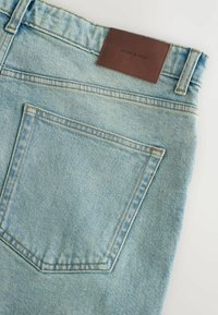 Heledad hall sinine teksapüksid, mille tagaküljel on kontrastsete õmblustega tasku ja pruuni nahklabeliga, millel on kirjas "DENIM SUPPLY" vöökoht.